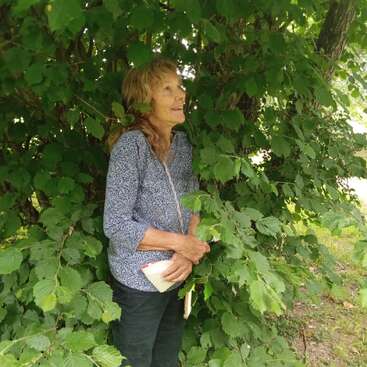 Una mujer está de pie bajo un árbol, rodeada de frondosas hojas verdes. Sonríe mientras sostiene un pequeño libro o cuaderno; lleva una blusa azul estampada y pantalones oscuros.