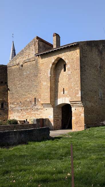 Esta imagen muestra un antiguo edificio de piedra con una entrada en arco, de arquitectura medieval, con un primer plano cubierto de hierba y el campanario de una iglesia visible al fondo, bajo un cielo azul y despejado.