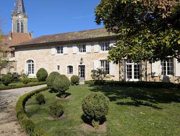 Una encantadora casa de piedra con contraventanas blancas y tejado de tejas, rodeada de arbustos bien cuidados, césped verde y árboles, con la torre de una iglesia visible en la distancia.