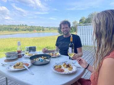 A man and woman are enjoying an outdoor meal with potatoes and wine, sitting at a table overlooking a serene lake and green countryside under a blue sky.