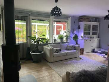 This cozy living room features a white sofa, lush green plants, large windows with garden views, a white cabinet, wood stove, and soft lighting, creating warmth.