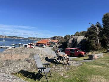 A man relaxes on a chair overlooking a scenic marina with sailboats. Red houses, a red van, rocky landscape, and clear blue skies complete the scene.