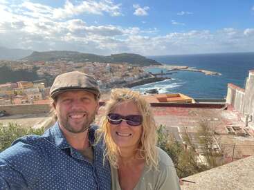 A smiling couple poses for a selfie with a coastal Mediterranean town, colorful buildings, green hills, and bright blue sea under a partly cloudy sky behind them.