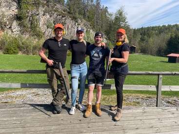 Four people pose outdoors on a wooden deck, holding rifles, wearing casual sporty clothing and caps. Green field and trees with rocky hillside in background.