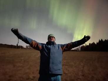 A person stands in an open field at night, arms outstretched, smiling under the beautiful green glow of the Northern Lights in the sky.