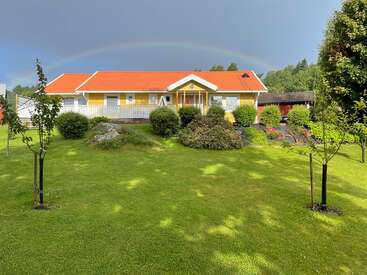 A charming yellow house with a red roof sits amid lush green lawns and gardens. A faint rainbow arches gently across the cloudy sky above.