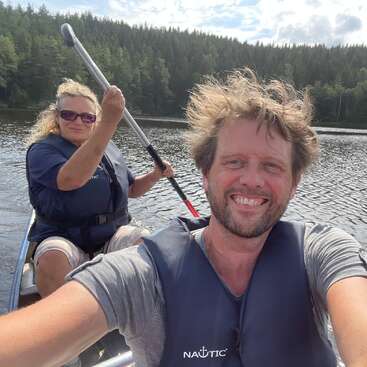 Ein lächelnder Mann und eine lächelnde Frau haben Spaß beim Kanufahren auf einem ruhigen See, umgeben von einem dichten Wald. Beide tragen Schwimmwesten und genießen diesen fröhlichen Moment im Freien.