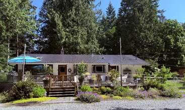 A cozy single-story house with a large wooden deck surrounded by lush greenery, colorful flowers, tall trees, outdoor seating, and a bright blue umbrella. Peaceful setting.