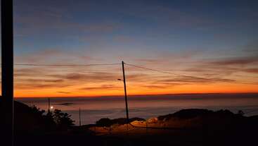 Um pôr do sol de tirar o fôlego sobre o mar, com tons vibrantes de laranja e azul se misturando no céu. Colinas em silhueta, um poste de luz e linhas de energia emolduram a cena tranquila.