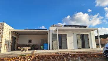 Uma casa moderna, de um andar, com painéis solares no telhado, é retratada sob um céu azul. Dois cães estão na frente, com materiais de construção por perto.