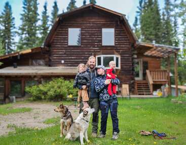 Eine glückliche Familie steht vor einem rustikalen Holzhaus, umgeben von grünem Gras und Bäumen. Zwei Erwachsene, zwei Kinder und zwei Hunde posieren gemeinsam und lächeln herzlich.