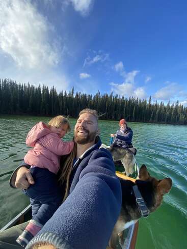 Ein lächelnder Mann macht ein Selfie in einem Kanu, zusammen mit einem kleinen Kind, einer Frau und einem Hund, auf einem ruhigen, grünen See, der von Kiefern umgeben ist.