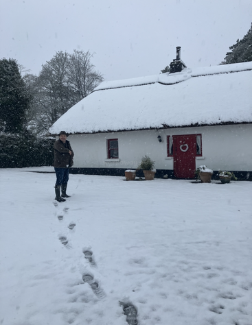 Uma pessoa está em meio à neve pesada do lado de fora de uma pequena cabana branca com telhado de palha, porta vermelha e vasos de plantas, cercada por árvores geladas. As pegadas levam você até lá.
