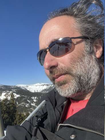 Ein Mann mit grauen Haaren und Bart trägt eine Sonnenbrille und eine schwarze Jacke und genießt einen sonnigen Tag in den verschneiten Bergen, hinter ihm erstreckt sich der blaue Himmel.