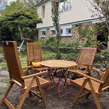 Cette image représente un coin de jardin accueillant, avec quatre chaises en bois et une table ronde en bois, disposées sur la terre. Une maison et de la verdure entourent cet espace.
