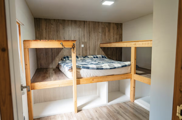 Um quarto aconchegante com beliches de madeira, roupa de cama xadrez, paredes com painéis de madeira e prateleiras de armazenamento abertas embaixo. Bem iluminado e minimalista, criando uma atmosfera rústica.