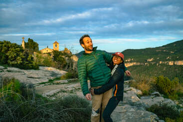 Um casal sorridente posa de forma divertida no topo de uma colina rochosa ao pôr do sol, cercado por vegetação, montanhas e uma pequena e antiga igreja de pedra à distância.