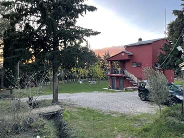 Uma casa vermelha de dois andares fica entre árvores altas ao pôr do sol. A entrada de automóveis de cascalho, o carro estacionado, o gramado verde e os assentos ao ar livre criam uma atmosfera rural e tranquila.
