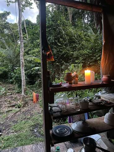 Une cuisine rustique en plein air donne sur une jungle verdoyante. Des étagères accueillent la vaisselle et les ustensiles, tandis qu'une bougie apporte une lumière chaleureuse. Le linge est étendu à l'extérieur, ce qui confère un charme particulier à la maison.