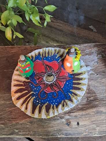 Une assiette décorative ronde ornée d'un soleil et d'une étoile vibrants contient deux porte-encens colorés émettant de la fumée, placés sur une surface en bois rustique à côté de feuilles de plantes vertes.