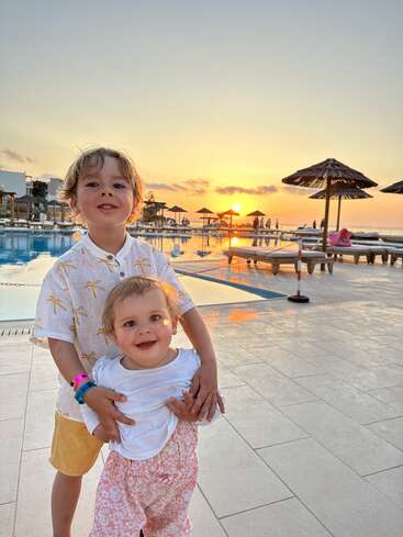 La imagen muestra a dos niños de pie en la terraza de una piscina al atardecer, con el niño abrazando a la niña y una serena piscina y el cielo de fondo.