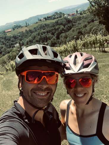 La imagen muestra un selfie de un hombre y una mujer en bicicleta, con casco y gafas de sol, frente a un paisaje de colinas y árboles.
