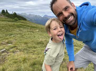 La imagen muestra a un hombre y un niño posando para una foto en un campo cubierto de hierba con montañas al fondo, ambos haciendo muecas.