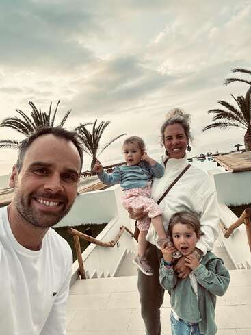 La imagen muestra a una familia de cuatro miembros posando para un selfie en una escalera blanca, con palmeras y un cielo nublado de fondo.