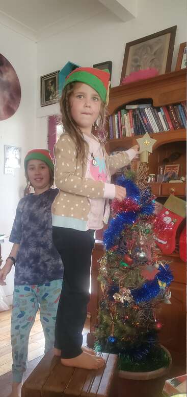 Duas crianças usando chapéus de duende decoram uma árvore de Natal com enfeites coloridos e guirlandas. Uma delas está em cima de um banquinho, pronta para colocar uma estrela no topo da árvore.