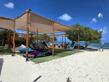 Eine sonnige Strandszene mit schattigen Holzdächern, Kitesurfing-Ausrüstung auf dem Sand, Bäumen, entspannenden Menschen, strahlend blauem Himmel, türkisfarbenem Meer und einem entfernten Drachen.