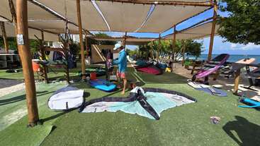Jemand steht im Schatten in Strandnähe, umgeben von Windsurf- und Kiteboard-Ausrüstung. Im Hintergrund sieht man das Meer und den blauen Himmel.