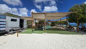 Ein Trainingsbereich am Strand mit schattigen Markisen, Picknicktischen, Fitnessgeräten und einem Anhänger liegt auf weißem Sand neben dem Meer unter strahlend blauem Himmel.