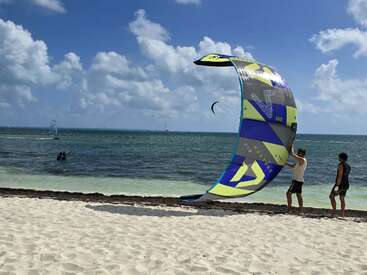 Zwei Leute auf einem Sandstrand machen einen bunten Drachen für's Kitesurfen fertig. Im Hintergrund sieht man die Wellen des Ozeans unter einem blauen Himmel mit flauschigen Wolken.