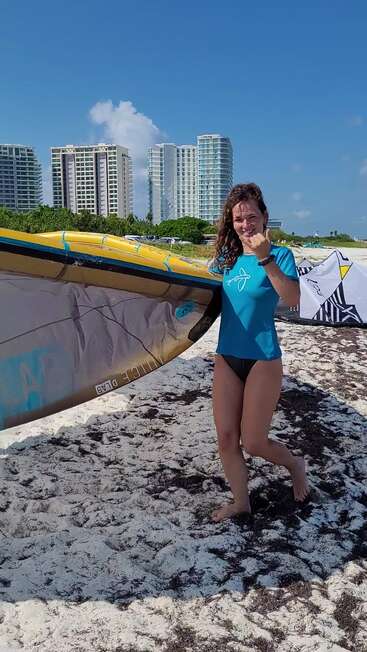 Uma mulher de camisa azul e biquíni preto carrega uma prancha de surfe numa praia arenosa, sorrindo, com prédios altos e um céu azul ao fundo.