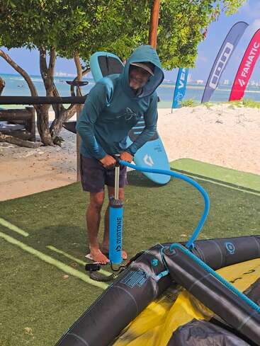 Um homem vestindo um moletom azul-petróleo está enchendo equipamentos infláveis para esportes aquáticos numa praia, com bandeiras coloridas, árvores e o mar visíveis ao fundo.