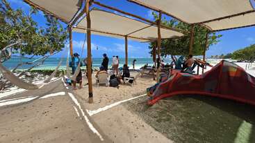 As pessoas relaxam sob toldos numa praia de areia, com redes, cadeiras e equipamentos de surfe por perto. O mar azul-turquesa e o céu azul criam uma atmosfera tropical.