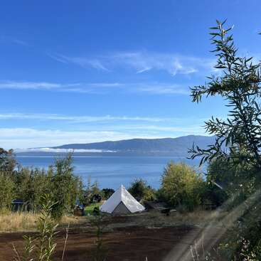 A white tent sits on a grassy hill overlooking a peaceful blue lake, surrounded by lush greenery, under a clear sky and distant mountains.