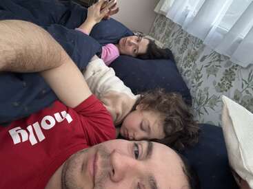 A cozy family scene in bed: a man in a red shirt, a sleeping child, and a woman lounging comfortably, using her phone under blue blankets.
