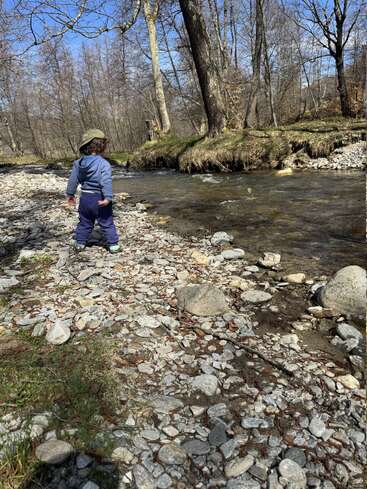 A young child in a blue outfit and green hat stands by a rocky riverside, surrounded by trees under a bright, clear blue sky. Peaceful nature scene.