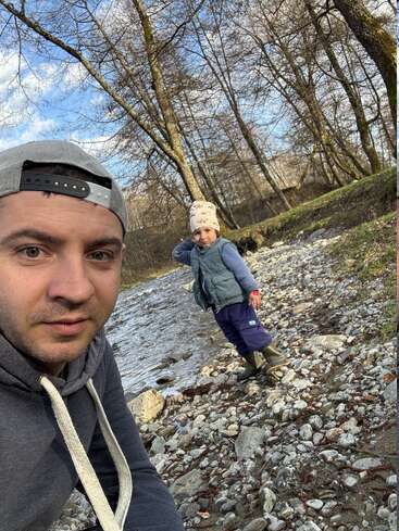 A man in a hoodie and backward cap takes a selfie outdoors, with a child standing on a rocky riverbank amidst leafless trees in early spring.