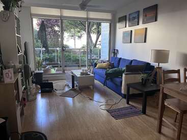 Uma sala de estar aconchegante com um sofá azul, piso de madeira, plantas de interior, mesa de centro, portas de pátio, luz natural, arte na parede e uma mesa de jantar. Atmosfera relaxante.
