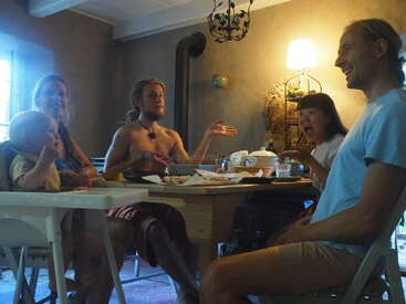 Cinco pessoas, incluindo um bebê, sentam-se ao redor de uma mesa de madeira e desfrutam de uma refeição em um ambiente fechado. A atmosfera é calorosa, aconchegante e cheia de risadas.