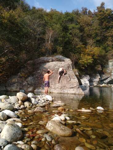Dos personas en la orilla rocosa de un río. Una está de pie mientras la otra salta al agua. A su alrededor hay piedras, árboles y un cielo azul despejado.