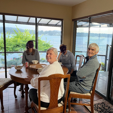 Four people are sitting around a wooden dining table, smiling. Behind them are large windows showing a beautiful lake view with trees and distant hills.