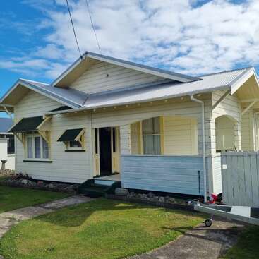 This image shows a charming, single-story, cream-colored house with green trim, a front porch, gable roof, manicured lawn, blue sky, and scattered white clouds.