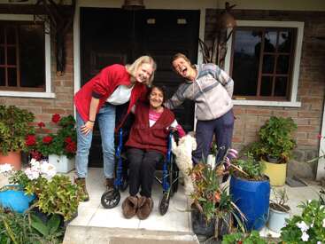 L'image montre trois femmes posant sous le porche d'une maison, avec une femme en fauteuil roulant et un chien blanc à ses côtés.