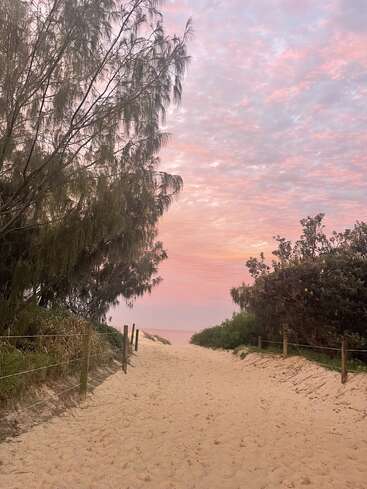 Um caminho de areia leva a uma vegetação exuberante em direção à praia. Árvores altas emolduram a cena sob um lindo céu rosa e roxo ao pôr do sol, criando um clima tranquilo.