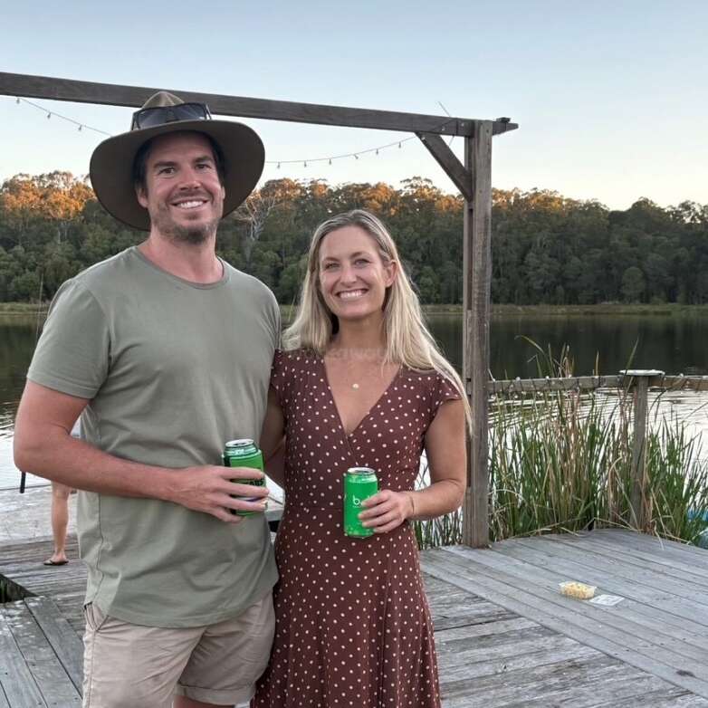 Um casal sorridente está em um deck de madeira à beira de um lago, segurando bebidas verdes. As árvores e a água criam um cenário tranquilo e natural. Luz do entardecer, clima relaxado.