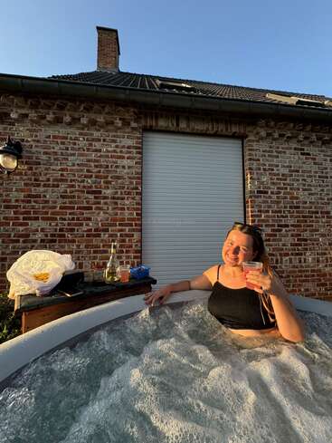 Una mujer se relaja en un burbujeante jacuzzi, con una copa en la mano y sonriendo. Detrás de ella hay una casa de ladrillo, bebidas, aperitivos y la luz del sol del atardecer.