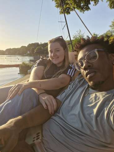 Una pareja sonriente se relaja en un muelle junto a un tranquilo lago al atardecer, disfrutando del ambiente tranquilo y cálido, rodeada de árboles y veleros.